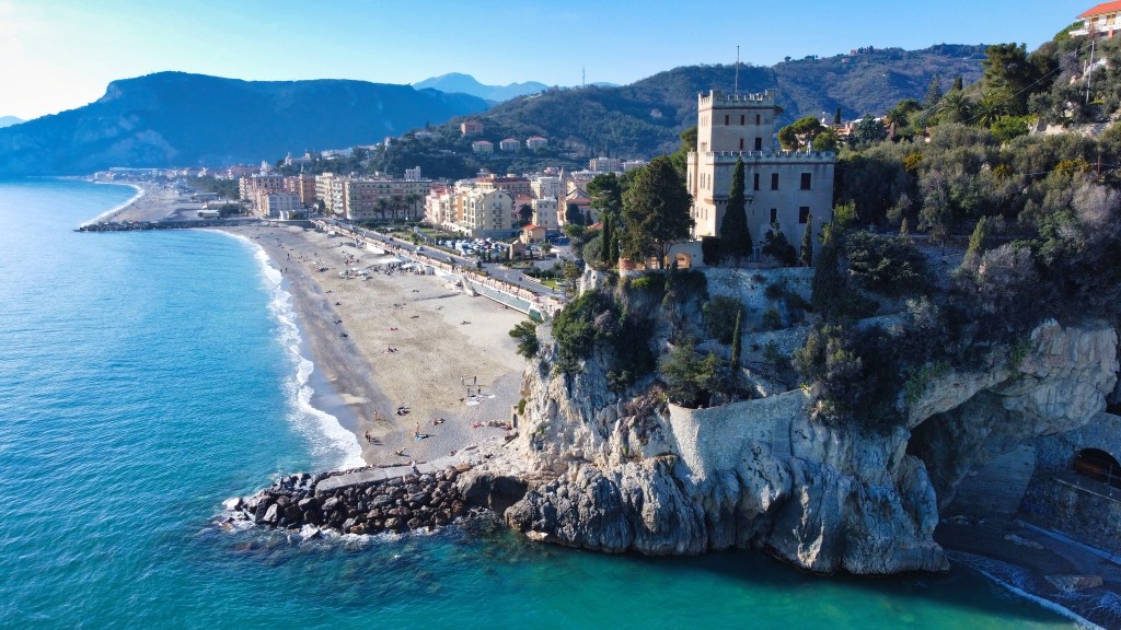 Immagine aerea di Finale Ligure mostrando il Castelletto su uno sperone roccioso affacciato sulla spiaggia e sul mare azzurro. Foto di Alessio Bevilacqua.