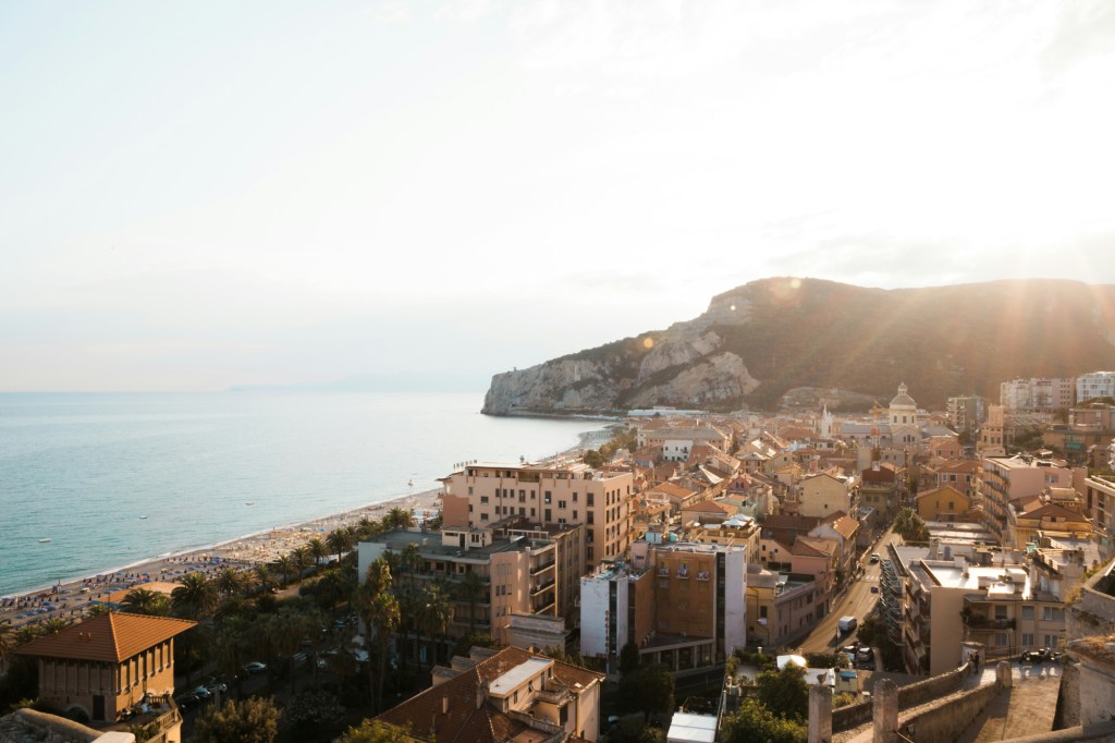 Una calda luce del tramonto illumina la città di Finale Ligure, mostrando il contrasto tra le antiche strutture e l'azzurro del mare nel background.